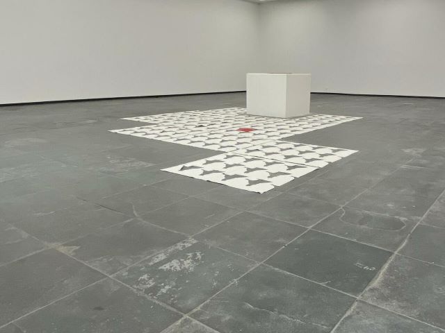 Ruimtelijke installatie met witte papieren vormen en een klein rood accent op een grijze museumvloer